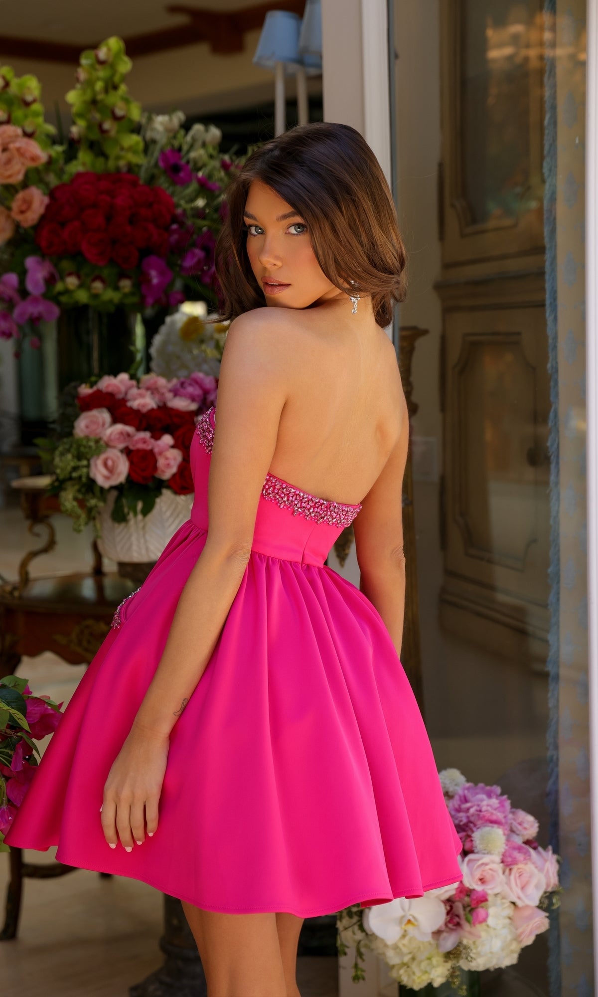 A woman indoors, surrounded by bouquets, looks over her shoulder while wearing the Ava Presley 29837 Strapless Short Hoco Dress in bright pink.